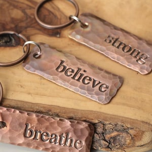 May include: Close-up of three copper keyrings with a hammered texture. The keyrings have the words "believe", "strong", and "breathe" stamped on them. Each keyring has a copper-coloured ring attached.