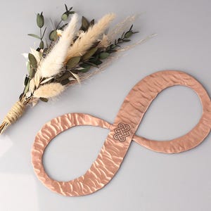 May include: A copper-colored infinity symbol with a textured surface and a Celtic knot design in the center. A small bouquet of dried flowers with green leaves and beige pampas grass is also in the image.