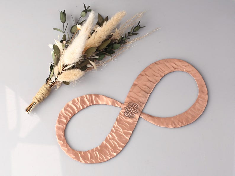 May include: A copper-colored infinity symbol with a textured surface and a Celtic knot design in the center. A small bouquet of dried flowers with green leaves and beige pampas grass is also in the image.