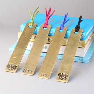 Pode incluir: Quatro marcadores de página retangulares de latão com textura martelada, cada um com um orifício no topo e um cordão de couro colorido em verde-limão, rosa choque, roxo ou preto. Os marcadores exibem símbolos gravados, incluindo nós celtas.