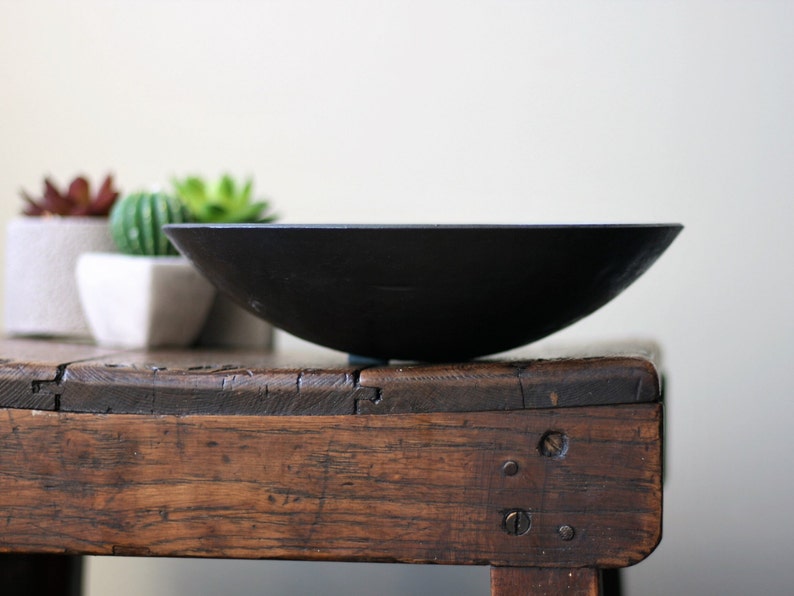 May include: A matte black, shallow, circular bowl with a smooth, curved exterior and a wide, open top. The bowl is constructed from a dark, solid material and features a simple, unadorned design with a consistent dark finish across its entire surface.