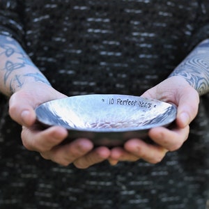 May include: A shallow, circular metal bowl with a hammered, textured surface. The interior rim is stamped with the text 10 Perfect Years, flanked by two small star symbols. The metal has a dark, brushed finish with visible indentations.