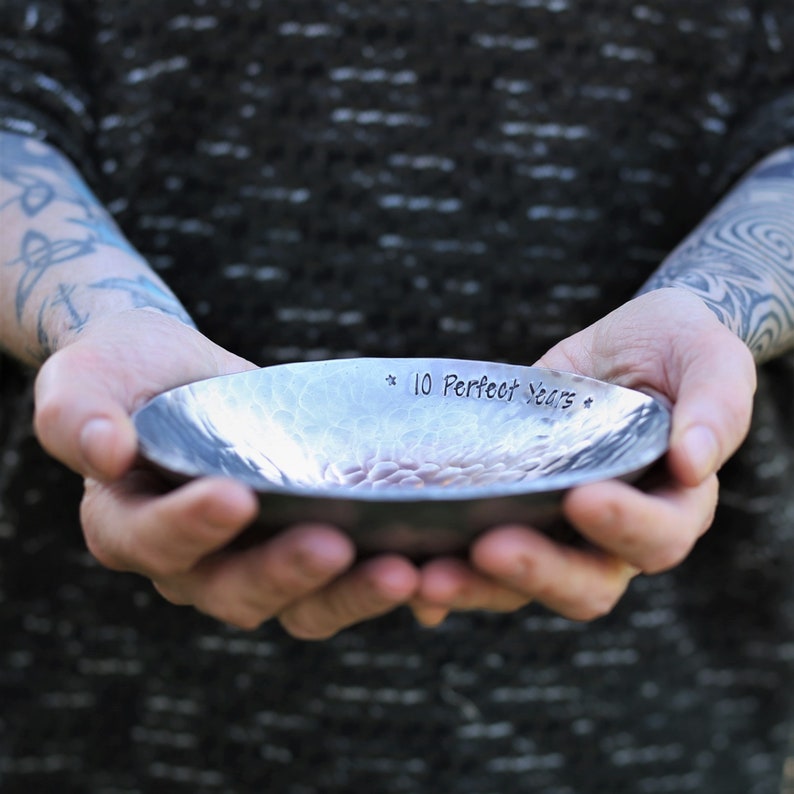 May include: A shallow, circular metal bowl with a hammered, textured surface. The interior rim is stamped with the text 10 Perfect Years, flanked by two small star symbols. The metal has a dark, brushed finish with visible indentations.