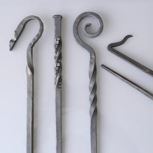 May include: Four handcrafted metal fireplace tools. The set includes a poker with a hook, a poker with a twisted design, a poker with a spiral design, and a small hook tool. The tools are made of a dark grey metal.