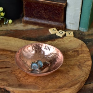 Peut inclure: Un petit bol rond en cuivre martel&eacute; avec le chiffre romain XXII estamp&eacute; sur le bord int&eacute;rieur. Le bol contient une paire de boutons de manchette argent&eacute;s aux faces circulaires bleu clair, pos&eacute; sur une surface en bois naturel.