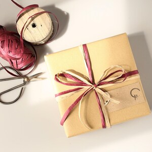 Puede incluir: Una caja de regalo cuadrada envuelta en papel marr&oacute;n, atada con un lazo de cinta de rafia color burdeos y crema. Se adjunta una peque&ntilde;a etiqueta rectangular de papel kraft con un logotipo circular negro. Cerca hay unas tijeras de metal y un carrete de rafia.