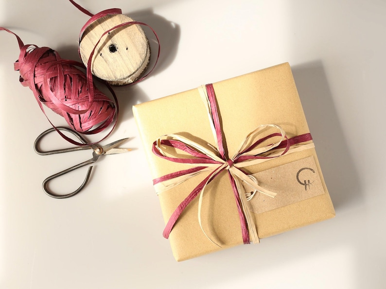 Puede incluir: Una caja de regalo cuadrada envuelta en papel marr&oacute;n, atada con un lazo de cinta de rafia color burdeos y crema. Se adjunta una peque&ntilde;a etiqueta rectangular de papel kraft con un logotipo circular negro. Cerca hay unas tijeras de metal y un carrete de rafia.