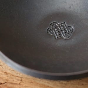 Può includere: Primo piano di una ciotola grigio scuro e poco profonda con un motivo a nodo celtico in rilievo al centro. La ciotola ha una finitura liscia e opaca e un bordo leggermente curvo. Lo sfondo è una superficie di legno.