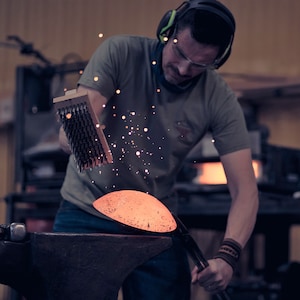 May include: A person wearing a green shirt and headphones uses a wire brush to clean a metal bowl. The bowl is sitting on an anvil.