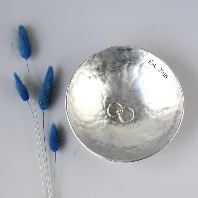 May include: A round, silver-coloured metal bowl with a hammered texture and the text Est. 2016 engraved on the inner rim. Two simple silver-coloured metal rings rest inside the bowl. Four blue dried grass stems are positioned to the left side.