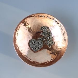 May include: A copper trinket dish with a hammered finish. The dish has the inscription "Every Year Better Than The Last" engraved on the rim. A silver heart-shaped pendant with a chain is resting in the dish.