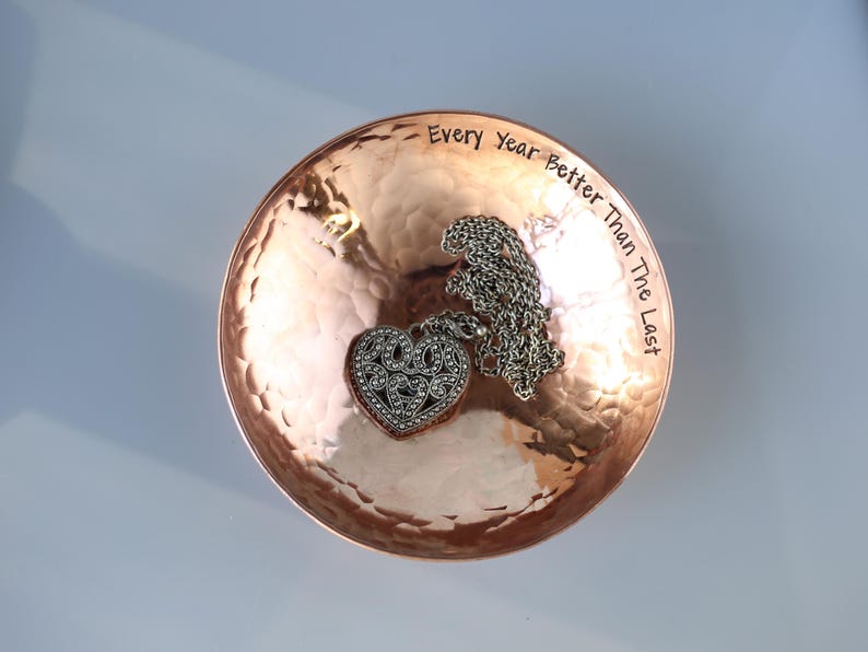 May include: A copper trinket dish with a hammered finish. The dish has the inscription "Every Year Better Than The Last" engraved on the rim. A silver heart-shaped pendant with a chain is resting in the dish.