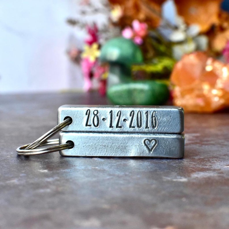 May include: A silver metal keychain with two rectangular tags. The top tag has the date "28-12-2016" engraved on it. The bottom tag has a small heart engraved on it.