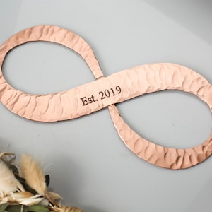 May include: Copper-colored infinity symbol with a hammered texture. The text "Est. 2019" is engraved on the center bar. The symbol is displayed against a gray background, with a floral arrangement in the lower left corner.