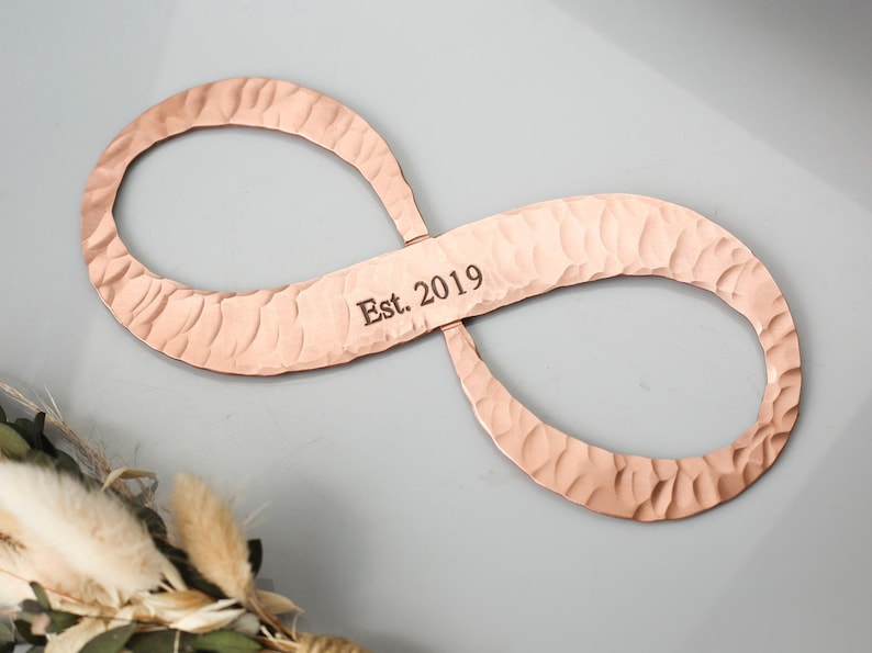May include: Copper-colored infinity symbol with a hammered texture. The text "Est. 2019" is engraved on the center bar. The symbol is displayed against a gray background, with a floral arrangement in the lower left corner.