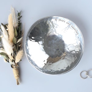 May include: A round, silver-toned hammered metal bowl with the text 10 years and counting engraved along the inner rim. Beside the bowl is a small bouquet of dried beige grasses and green eucalyptus leaves, along with two simple silver rings.
