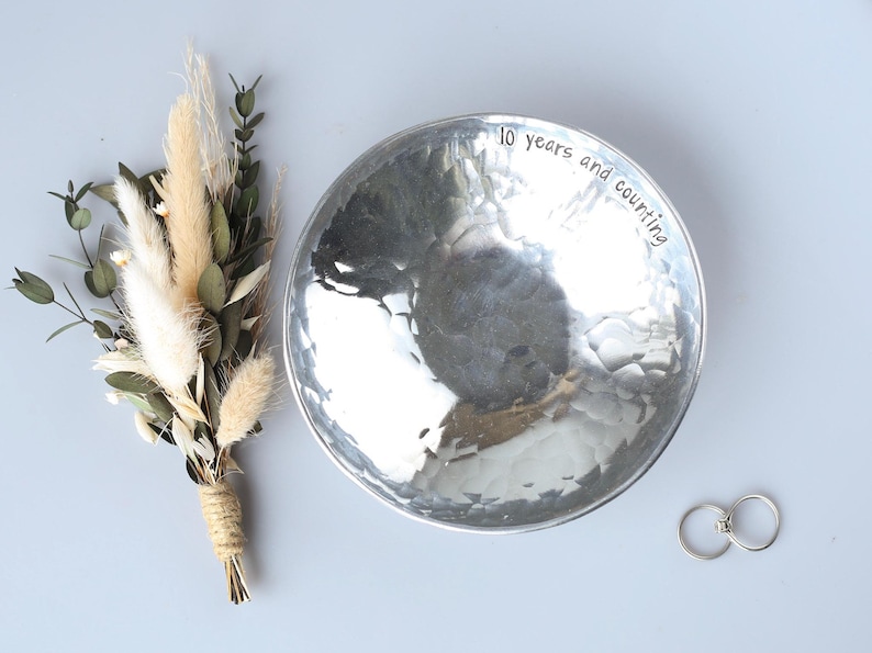 May include: A round, silver-toned hammered metal bowl with the text 10 years and counting engraved along the inner rim. Beside the bowl is a small bouquet of dried beige grasses and green eucalyptus leaves, along with two simple silver rings.