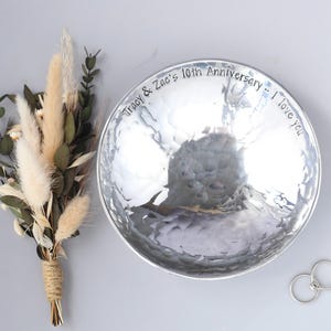 May include: A round, hammered silver-toned metal bowl with the engraved text "Tracy & Zac's 10th Anniversary -- I love you" along the inner rim, accompanied by a small bouquet of dried beige grasses and green leaves, and two silver rings.