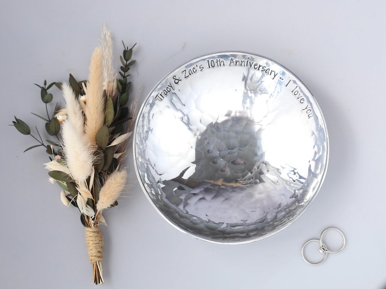 May include: A round, hammered silver-toned metal bowl with the engraved text "Tracy & Zac's 10th Anniversary -- I love you" along the inner rim, accompanied by a small bouquet of dried beige grasses and green leaves, and two silver rings.