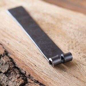 May include: A hand-forged, dark metal object with a flat, rectangular body and two cylindrical, hollow ends. The metal has a textured, matte finish. The object rests on a light brown, textured wooden surface.