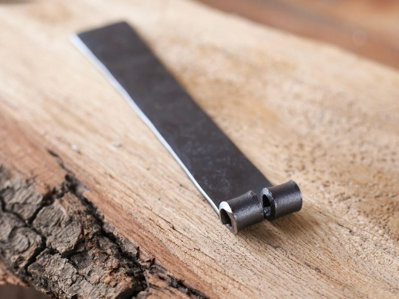 May include: A hand-forged, dark metal object with a flat, rectangular body and two cylindrical, hollow ends. The metal has a textured, matte finish. The object rests on a light brown, textured wooden surface.