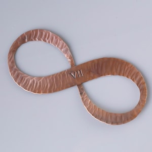 May include: A copper-colored infinity symbol with a hammered texture. The Roman numeral "VIII" is stamped in the center. The symbol is displayed on a light gray background.
