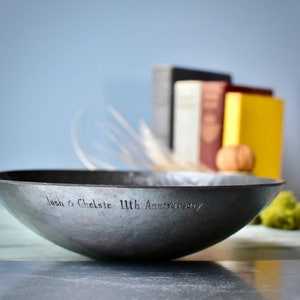 May include: A dark grey, shallow metal bowl with a hammered texture. The exterior rim is engraved with the text "Josh & Chelsie 11th Anniversary" in a simple, sans-serif font. The bowl has a wide, open circular shape with a smooth interior.
