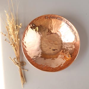 May include: A round, hammered copper bowl with a polished, reflective surface. The inner rim is engraved with the text 26 June 2004 - Something Changed. Beside the bowl lies a small, tied bundle of dried, light-tan wheat stalks and grasses.