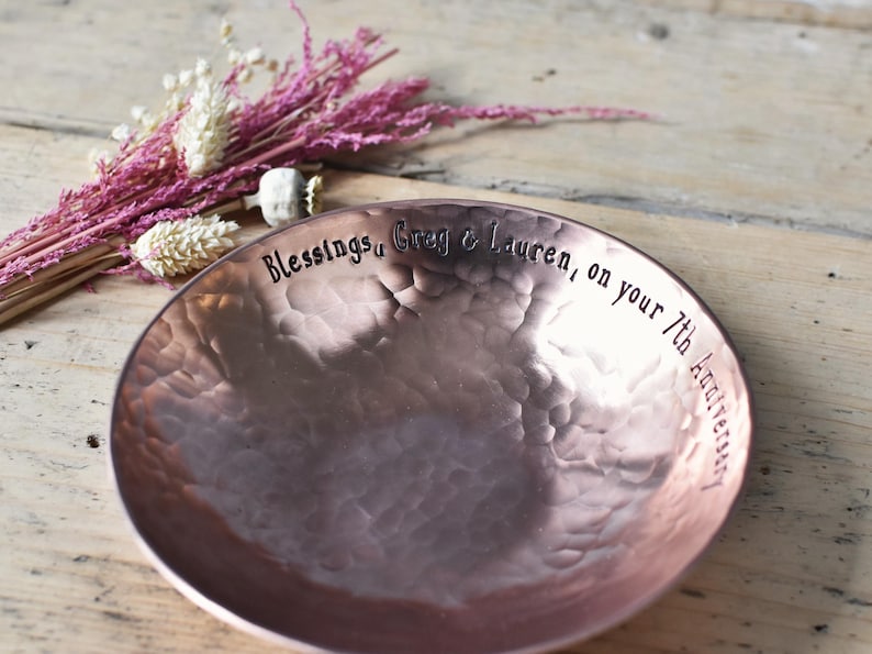 A bright hammered copper bowl with an inscription on the inside rim that reads, 'Blessings, Greg & Lauren, on your 7th Anniversary'. The hand crafted bowl sits on a wooden slatted table next to a small pink posy of dried flowers.