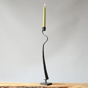 Op de afbeelding: Een lange, zwarte metalen kandelaar met een slank, gebogen ontwerp. Hij houdt een brandende, lichtgroene kaars vast. De basis is vierkant en de kandelaar heeft een unieke, artistieke vorm. De kaars is ongeveer 25 cm hoog.
