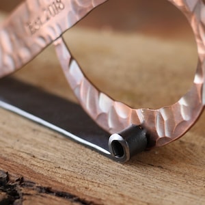 May include: Close-up of a copper bottle opener with a textured surface and a dark metal handle. The text "Est. 2018" is stamped on the copper. The opener is on a wooden surface.