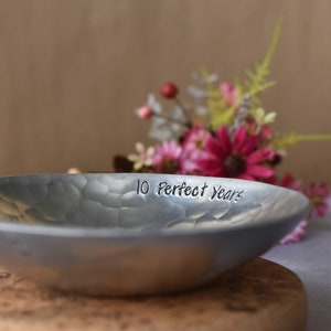 May include: A shallow, circular metal bowl with a hammered, textured surface. The interior rim of the bowl is engraved with the black text '10 Perfect Years' in a simple, sans-serif font. The metal has a matte, silver-toned finish.