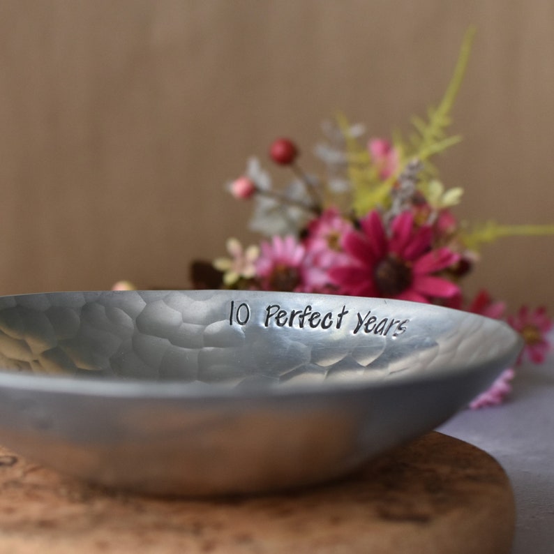 May include: A shallow, circular metal bowl with a hammered, textured surface. The interior rim of the bowl is engraved with the black text '10 Perfect Years' in a simple, sans-serif font. The metal has a matte, silver-toned finish.