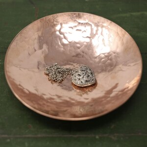 Peut inclure: Un bol rond en cuivre martelé avec une finition métallique réfléchissante. À l'intérieur du bol repose un collier argenté doté d'un pendentif en forme de cœur orné de petites pierres claires et scintillantes disposées en un motif complexe.