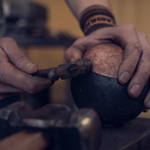 Peut inclure: Un petit marteau en m&eacute;tal avec un manche en bois est utilis&eacute; pour fa&ccedil;onner une sph&egrave;re en cuivre textur&eacute;e reposant sur une base m&eacute;tallique sombre et arrondie. La surface en cuivre pr&eacute;sente des marques de martelage, un autre marteau est visible.