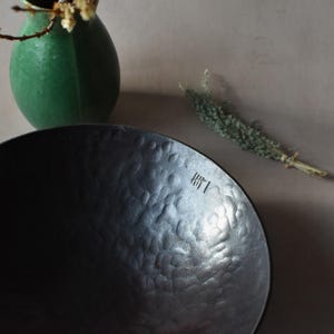 May include: A dark charcoal-coloured bowl with a hammered, dimpled texture on its interior surface. The bowl features a small, stamped mark consisting of vertical lines near the rim. The finish is matte and dark, contrasting with the light-coloured background.