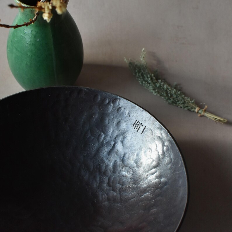May include: A dark charcoal-coloured bowl with a hammered, dimpled texture on its interior surface. The bowl features a small, stamped mark consisting of vertical lines near the rim. The finish is matte and dark, contrasting with the light-coloured background.