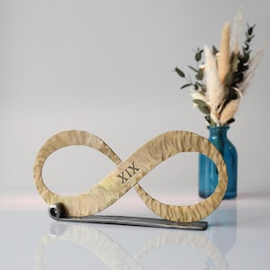May include: A metal infinity symbol with a textured, gold-toned finish, featuring the Roman numeral "XIX". The symbol rests on a dark grey base. A blue vase with dried flowers is in the background.