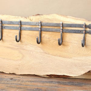 Puede incluir: Un perchero rústico de metal forjado a mano con cinco ganchos curvos. El perchero está montado sobre una tabla de madera natural, mostrando la veta y la textura de la madera. El metal tiene un acabado oscuro y envejecido.