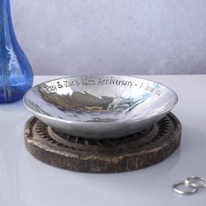 May include: A shallow, hammered silver-toned metal bowl with the engraved text 'Tracy & Zac's 10th Anniversary - I love you' along the inner rim. The bowl rests on a dark, circular, textured base, accompanied by two silver rings nearby.