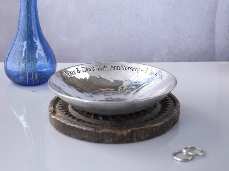 May include: A shallow, hammered silver-toned metal bowl with the engraved text 'Tracy & Zac's 10th Anniversary - I love you' along the inner rim. The bowl rests on a dark, circular, textured base, accompanied by two silver rings nearby.