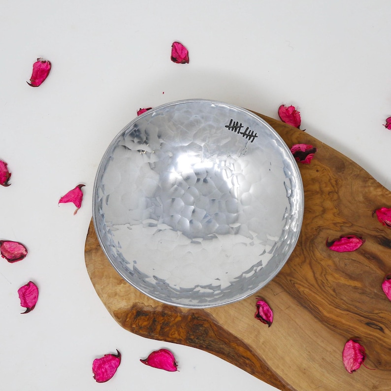 May include: A round, silver-coloured metal bowl with a hammered, textured surface. The interior rim features a small, black, hand-drawn tally mark design consisting of five vertical lines crossed by a diagonal line, indicating the number five.