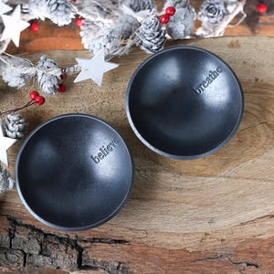 May include: Two small, dark grey, round bowls with the words "believe" and "breathe" embossed on the inside. The bowls are on a wooden surface, decorated with a garland of white stars, pinecones, and red berries.