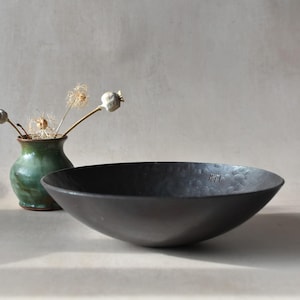 May include: A dark charcoal grey, shallow, hammered metal bowl with a textured surface and a small stamped mark on the interior rim, positioned next to a small, green, glazed ceramic vase containing several dried poppy seed pods.