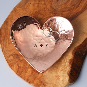 May include: A heart-shaped copper trinket dish with a hammered texture, featuring the stamped letters A + Z in the centre. Two silver-toned metal rings rest inside the dish, which is placed on a piece of natural wood with a live edge.