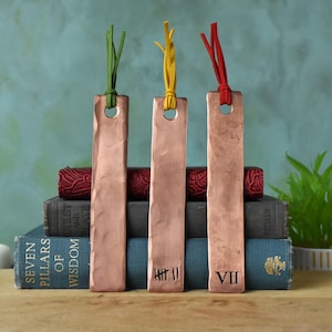 May include: Three copper bookmarks with leather tassels. The bookmarks are engraved with the numbers "VII", "III", and "VII". The bookmarks are on top of a stack of books.