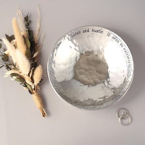 May include: A round, silver-coloured hammered metal bowl with the engraved text "Sarah and Austin 10 a&ntilde;os de matrimonio" along the inner rim. Beside the bowl is a small bouquet of dried beige grasses and green leaves, plus two silver rings.