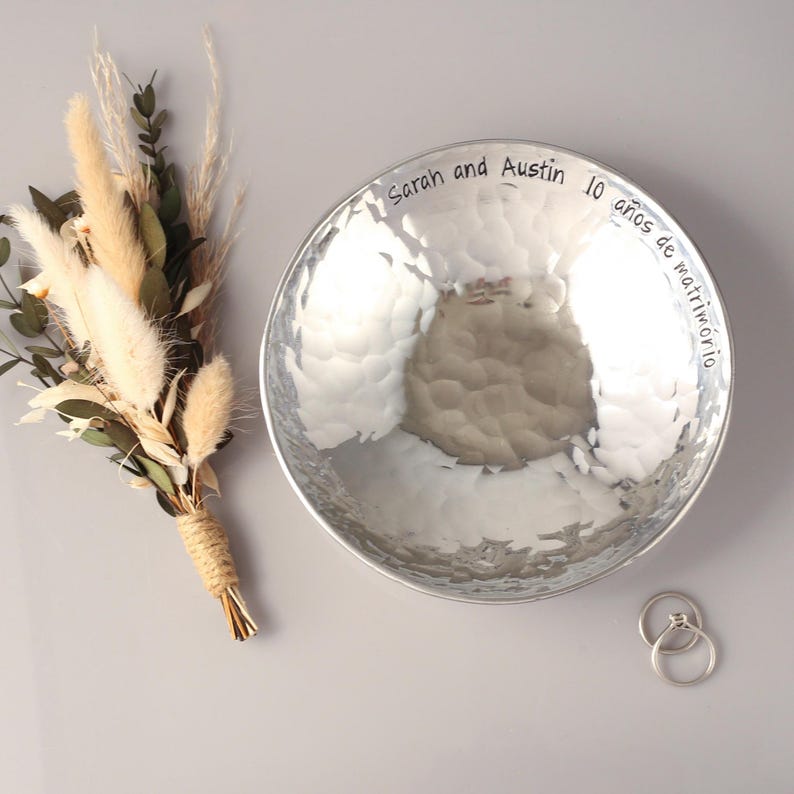 May include: A round, silver-coloured hammered metal bowl with the engraved text "Sarah and Austin 10 a&ntilde;os de matrimonio" along the inner rim. Beside the bowl is a small bouquet of dried beige grasses and green leaves, plus two silver rings.