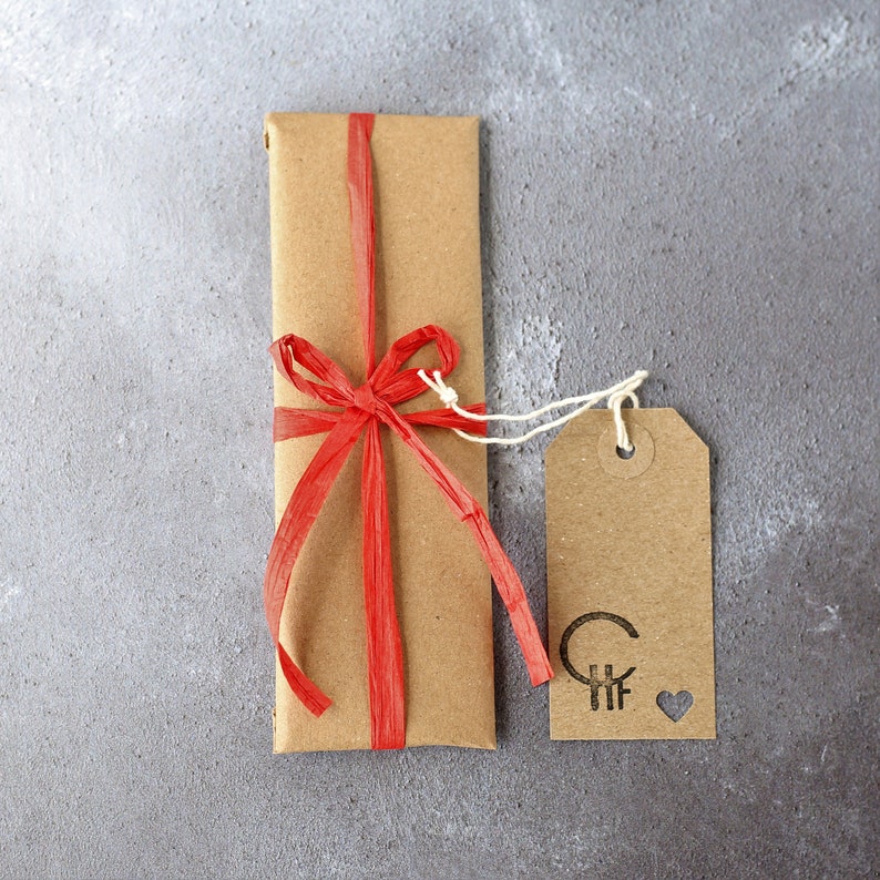 Pu&ograve; includere: Una busta regalo di carta marrone con un nastro e un fiocco rosso. Un'etichetta marrone con il testo "CHF" e un cuore &egrave; attaccata alla busta.