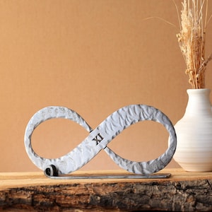 Può includere: Scultura a forma di simbolo dell'infinito argentato, con il numero romano XI sul lato sinistro. La scultura strutturata è posta su una superficie di legno. Un vaso bianco con fiori secchi è a destra.